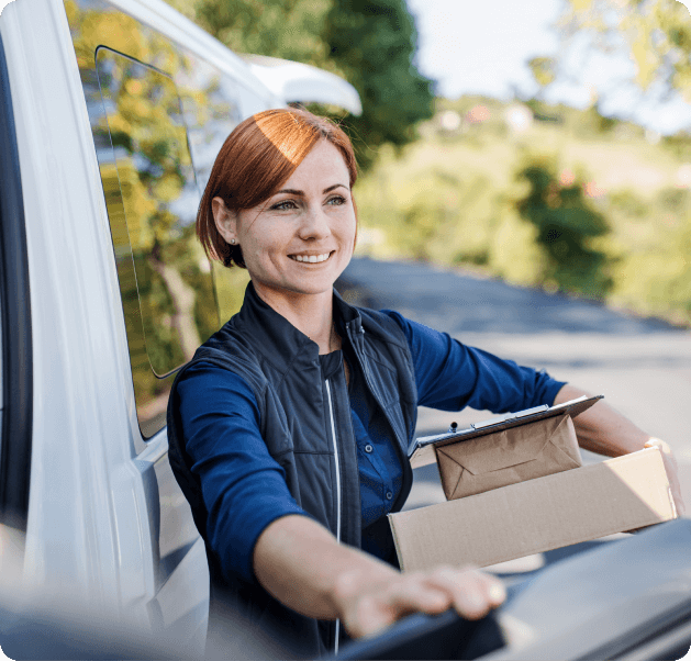 A smiling, female delivery driver exiting a vehicle while holding a stack of boxes. 